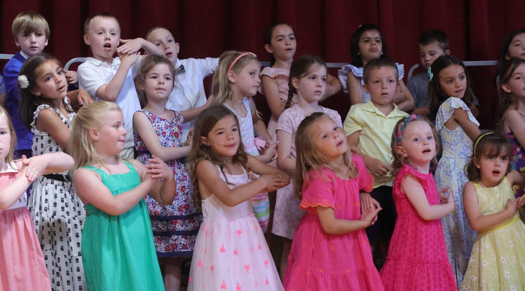 Group of kindergarten children wearing dressy clothes perform on risers during kindergarten graduation.