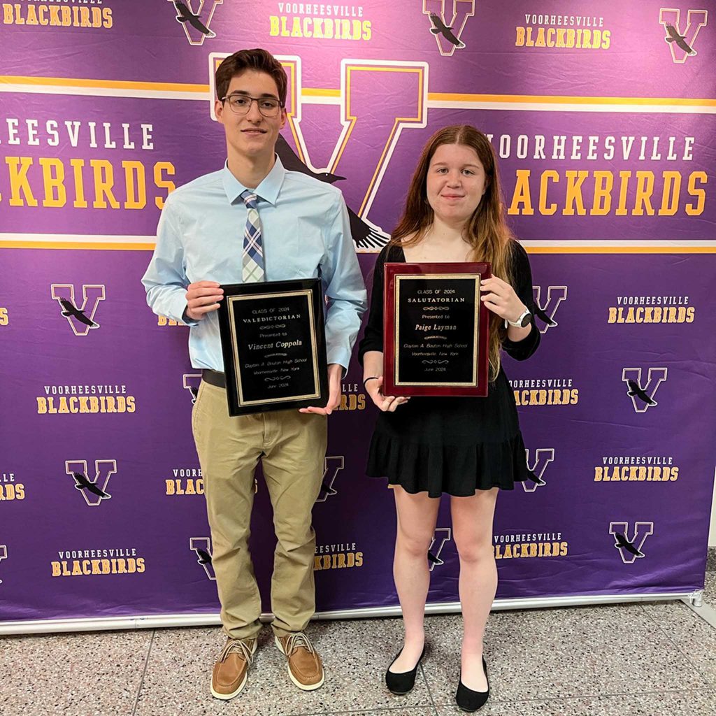 Valedictorian and salutatorian stand infront of Voorheesville Blackbird backdrop. They are dressed in professional attire and each is holding a plaque denoting their academic achievement.