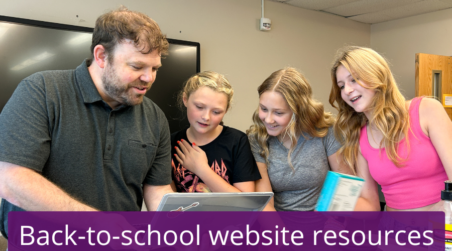 A teacher and group of high school age students gather around a laptop to view its screen in a high school classroom. 