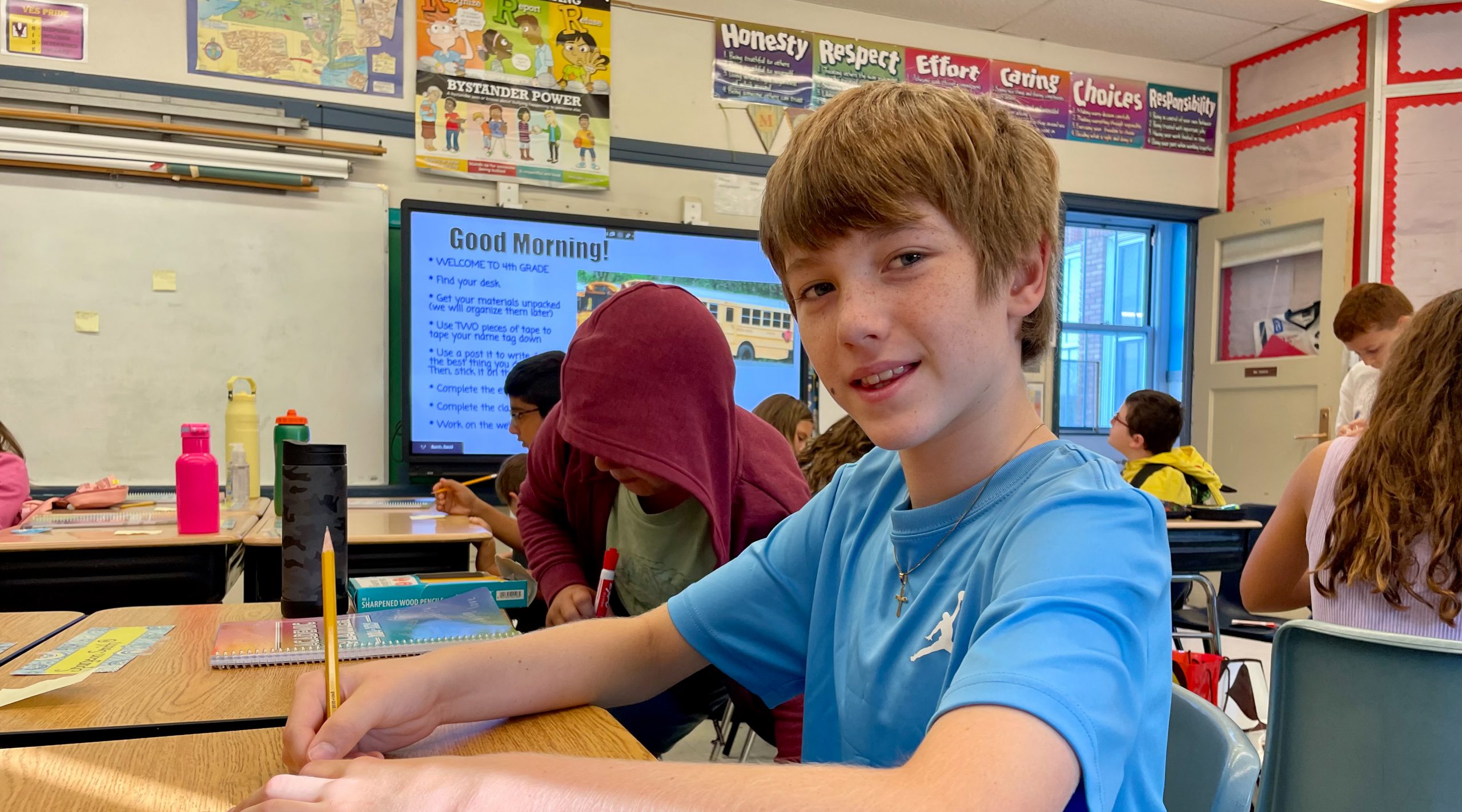 Boy sits in classroom with activity going on adjacent to him. Classroom walls are decorated with learning materials. He is smiling.