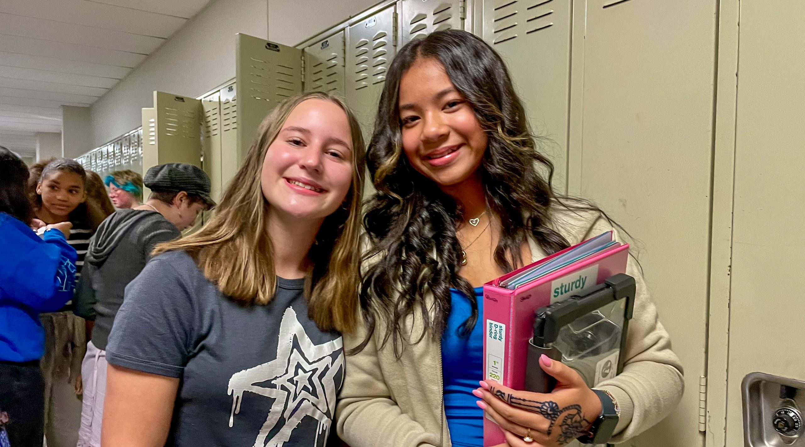 Two middle school girls stand in front of lockers. They are smiling. One is holding books and a binder.