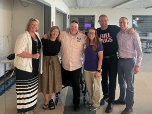 Six adults stand with arms around each other in cafeteria. One is wearing a shirt that says "free hugs." 