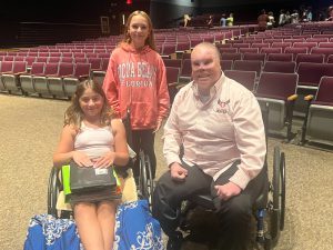 Two middle school students, one in a wheelchair are next to a man in a wheelchair.