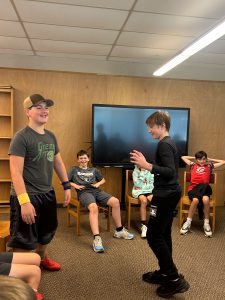 Two boys stand with other students sitting near by. They are smiling and participating in circle activity.