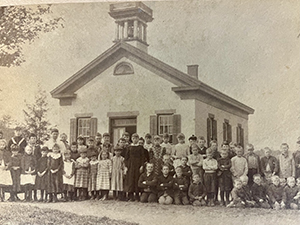 A sepia toned photo featuring an old fashioned school building with a group of students gathered for a photo in front of it. This is the first school building for Voorheesville students.