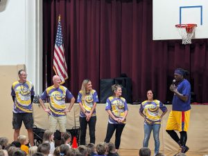 Five adults in matching shirts stand next to a member of the Harlem Wizards at an assembly.