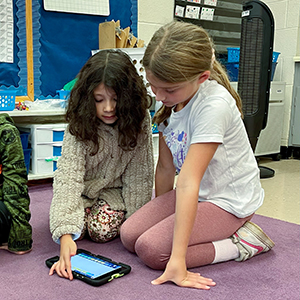 Two young students kneel together on a classroom carpet and work on a lesson on a computer tablet. 