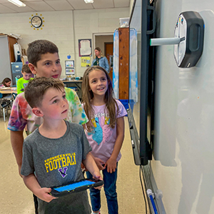 Three young students gather together at a classroom whiteboard. The student at the front is holding a tablet that is being used to move an iRobot up and down on the wall board. 