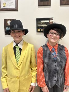 Two students stand together. They are dressed in suits and ties and are wearing hats.