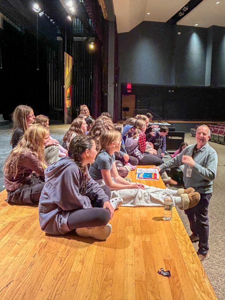 Students sit cross-legged on stage and receive direction from Steve Suriano. Many are smiling.