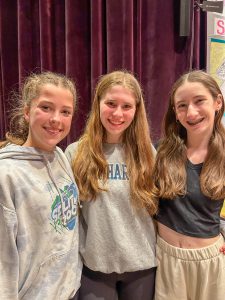 Three high school students stand with arms around each other. Stage curtain in background.