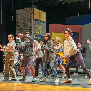 Group of students, some wearing hats and microphones dance on stage. 