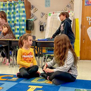 Two girls sitting on carpet face each other. Other students in background of classroom.