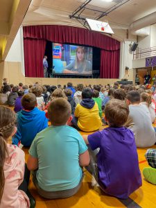 Students sit on floor for an assembly. Large screen on stage shows a child speaking.