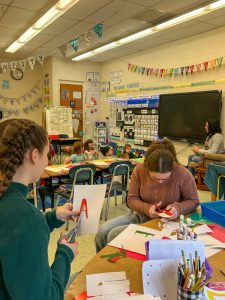 In foreground, two teens cut materials for a craft project. In background, teacher gives a lesson to grade 1 students as they sit on the floor.