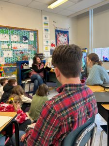 Two teens look on as teacher reads a book to a group of young students sitting on a carpet.