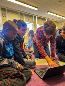 Teen on floor with grade five students. They are clustered around a laptop computer.