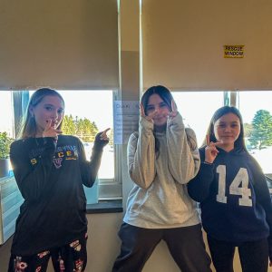 Three students stand in front of a clue taped to a wall. They are making peace gestures with their hands.