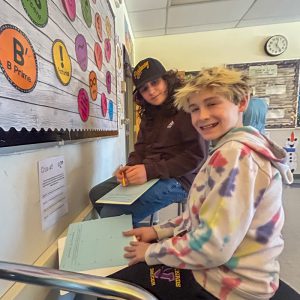 Two students stand in front of clue. They have papers they are writing on.