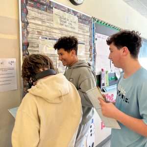 Three students stand in front of clue. They have papers they are writing on.