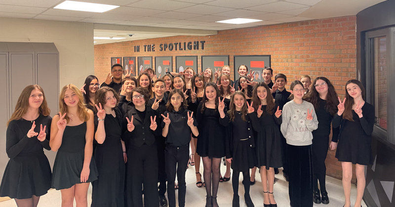 Dressed in concert black, members of middle school choral group stand together in hallway. They are giving the peace sign.